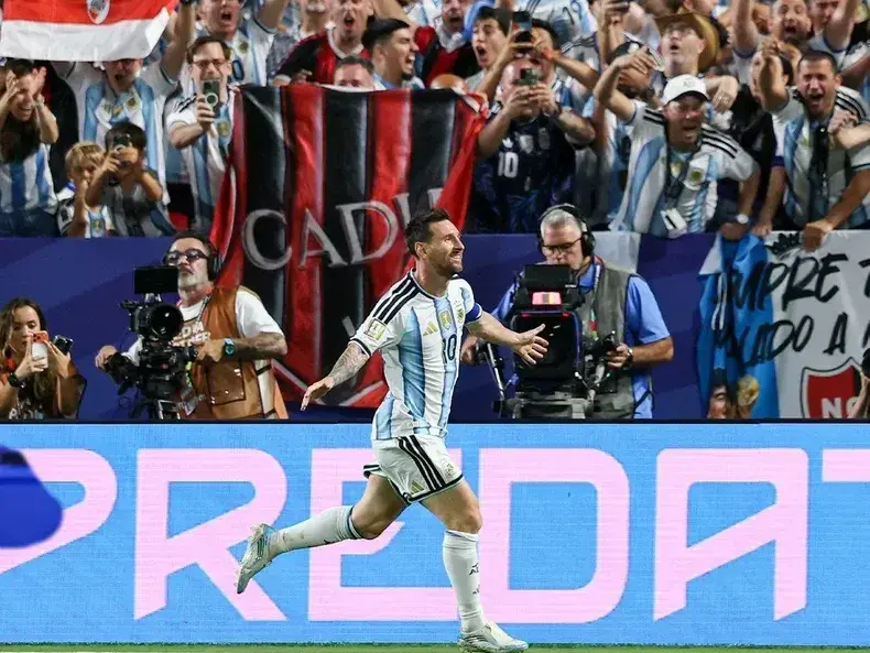 Lionel Messi festejando uno de sus tres goles y detrás, en las gradas, la bandera del fogonero de Pergamino.