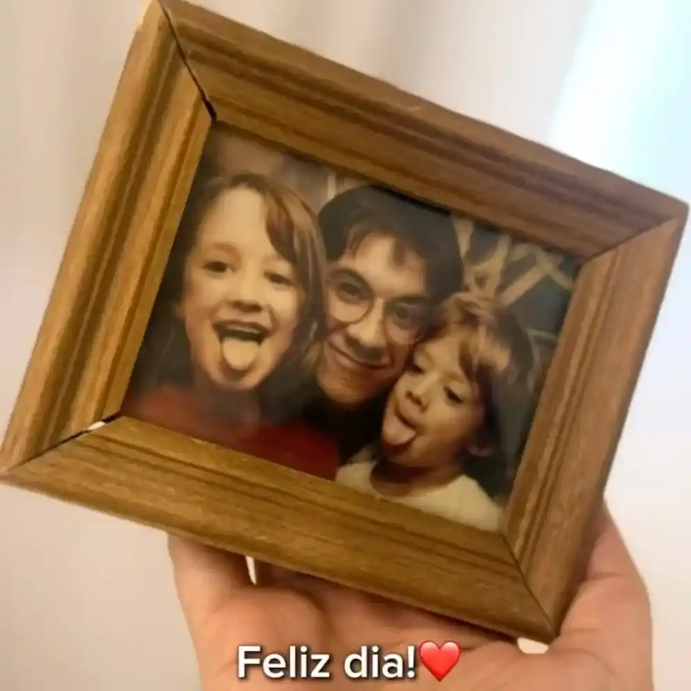 Una mano sostiene un marco de madera que contiene una fotografía en blanco y negro donde Piñón Fijo aparece sin maquillaje junto a sus dos hijos, imagen difundida con motivo del Día del Padre.
