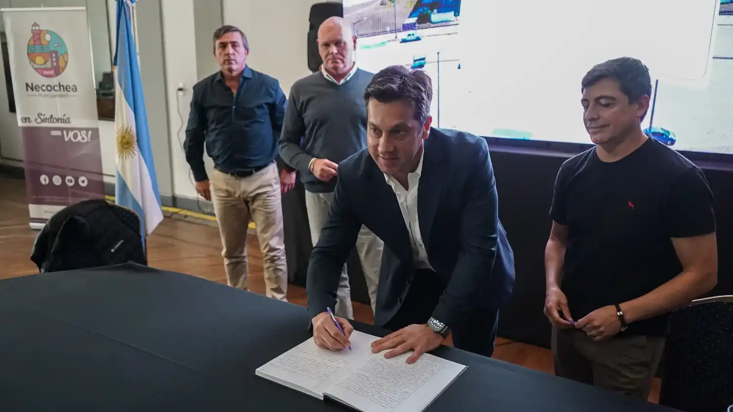 Firma. Arturo Rojas firma el acta, y a su lado Oscar Merlo de A Toda Vela Mar S.A. Más atrás Gertie y Furno observan la escena.